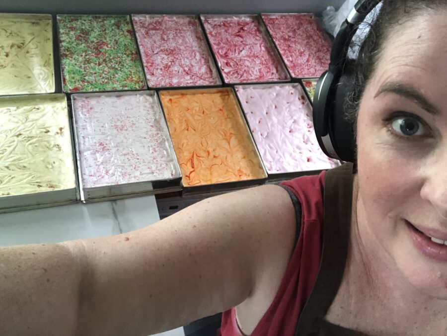 9 different colour trays of marshmallows with Paula to the right of the image, wearing headphones, as a selfie photo.